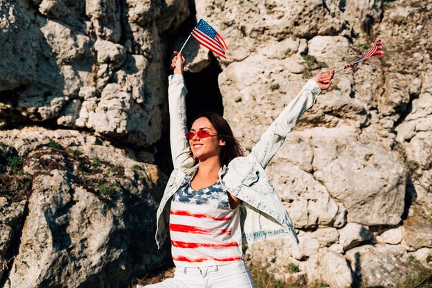 Mujer joven alegre que agita banderas americanas en rocas