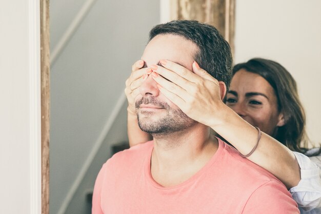 Mujer joven alegre que cubre los ojos de su novio con las manos y lo lleva a su nuevo apartamento.