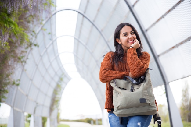 Mujer joven alegre que se inclina en el bolso en parque de la ciudad