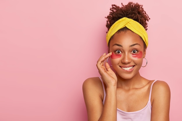 Mujer joven alegre que muerde los labios, tiene una expresión feliz, una piel de cara sana, usa parches debajo de los ojos para reducir las bolsas, tiene procedimientos antiarrugas