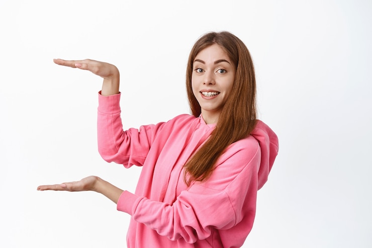Foto gratuito mujer joven alegre que muestra gran tamaño grande en copyspace, producto de exhibición o logotipo, sosteniendo un espacio vacío para pancarta, de pie sobre una pared blanca
