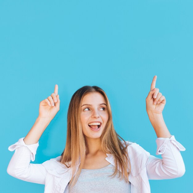 Mujer joven alegre que señala su dedo hacia arriba contra fondo azul