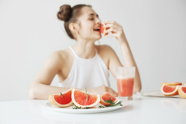 Mujer joven alegre que sonríe sentado en la tabla que come el pedazo de pomelo sobre la pared blanca. Concepto de comida saludable.