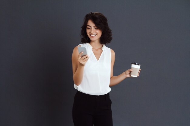 Mujer joven alegre que sostiene su teléfono y café en manos
