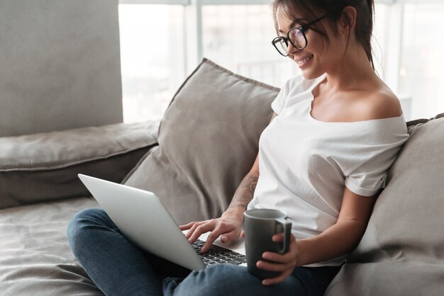 Mujer joven alegre que sostiene la taza de café usando la computadora portátil.