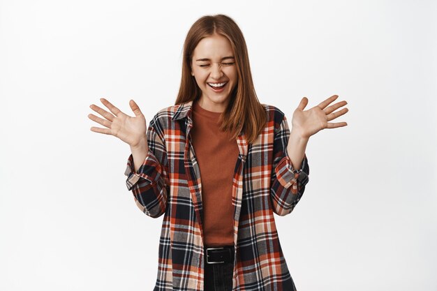 Mujer joven alegre riendo, saltando de la felicidad y celebrando la victoria, gritando de alegría, dándose la mano, de pie contra la pared blanca emocionada.