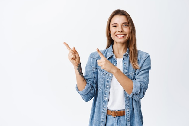 Mujer joven alegre en ropa casual mostrando publicidad, señalando con el dedo en la esquina superior izquierda y sonriendo, recomendando oferta, de pie en blanco