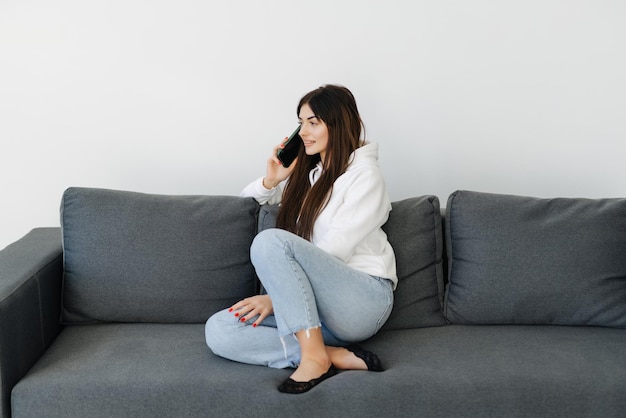 Mujer joven alegre sentada en un sofá en casa hablando por teléfono móvil