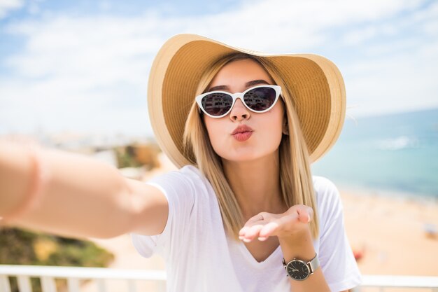 Mujer joven alegre con sombrero y gafas de sol envía besos tomando selfie con teléfono móvil en verano