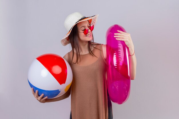 Mujer joven alegre con sombrero con gafas de sol rojas sosteniendo una bola inflable y un anillo sonriendo mirando el anillo inflable de pie en blanco