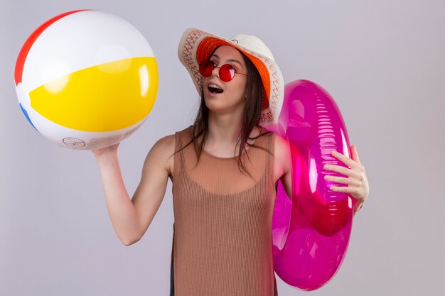 Mujer joven alegre con sombrero con gafas de sol rojas sosteniendo bola inflable y anillo sonriendo de pie sobre blanco