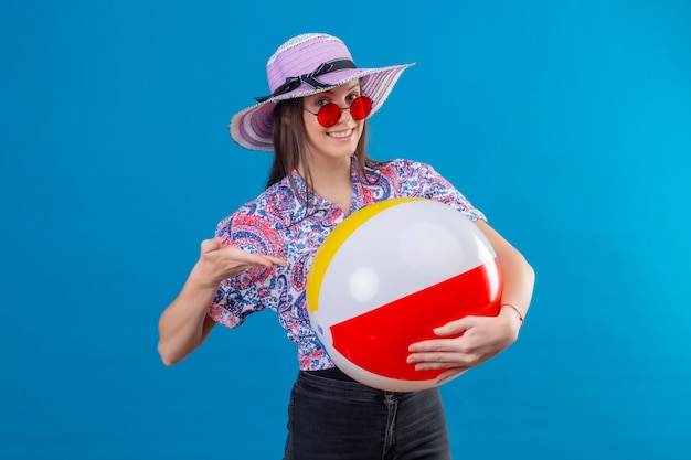 Foto gratuita mujer joven alegre con sombrero con gafas de sol rojas sosteniendo una bola inflable apuntando con el brazo de la mano a ella sonriendo con cara feliz de pie en azul