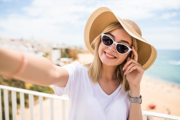 Mujer joven alegre con sombrero y gafas de sol tomando selfie con teléfono móvil en el mar de resort de verano