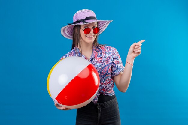 Mujer joven alegre con sombrero de verano con gafas de sol rojas sosteniendo una bola inflable apuntando con el dedo hacia el lado sonriendo con cara feliz de pie sobre el espacio azul
