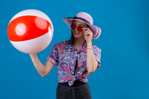 Mujer joven alegre con sombrero de verano con gafas de sol rojas sosteniendo una bola inflable mirando a un lado con una sonrisa feliz de pie sobre el espacio azul