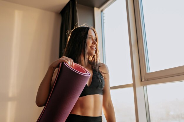 Mujer joven alegre sosteniendo colchoneta de yoga contra un fondo claro Morena de pelo largo en las mejores sonrisas mostrando sus dientes y mirando la ventana Concepto de fitness en el hogar