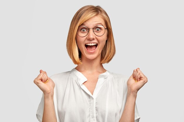 La mujer joven alegre tiene una expresión positiva, aprieta los puños, tiene una mirada llena de alegría, está muy animada, viste una camisa blanca, aislada sobre una pared blanca. Gente, felicidad, concepto de éxito