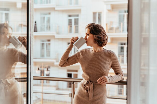 Mujer joven alegre tomando café y mirando la ciudad. Foto interior de niña rizada complacida en vestido marrón pasando la mañana en el balcón.