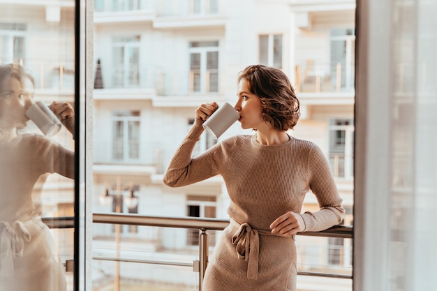 Mujer joven alegre tomando café y mirando la ciudad. Foto interior de niña rizada complacida en vestido marrón pasando la mañana en el balcón.