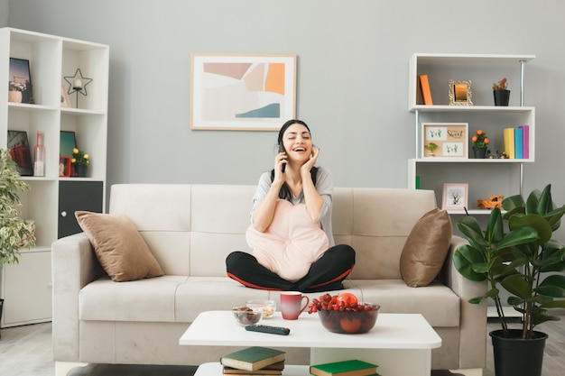 Foto gratuita mujer joven con almohada sentada en el sofá detrás de la mesa de café habla por teléfono en la sala de estar