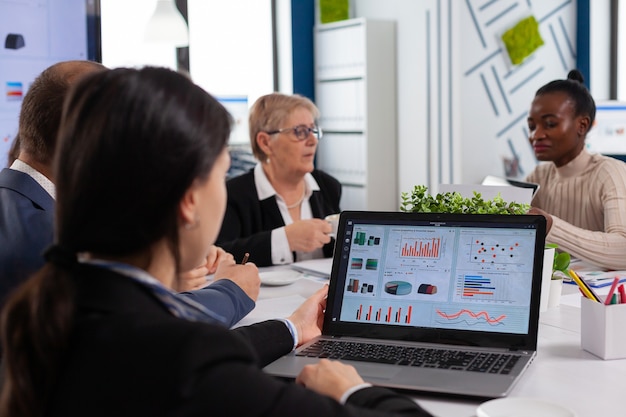 Mujer joven analizar gráficos en la computadora portátil en la puesta en marcha de la sala de reuniones de negocios