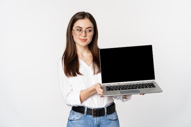 Mujer joven con anteojos que muestra la pantalla de una computadora portátil, demostrando promoción en la computadora, sitio web o tienda, de pie sobre fondo blanco