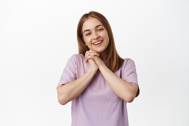 Mujer joven apasionada admira algo hermoso, mirando con fascinación, ojos románticos de simpatía, enamorarse, sonriendo y mirando al frente, de pie contra la pared blanca