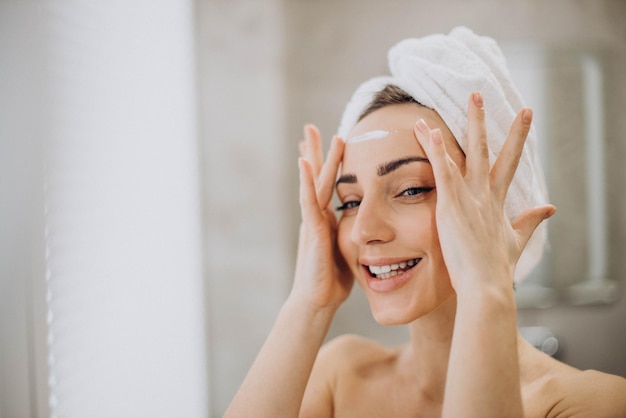 Mujer joven aplicando crema facial en la cara con una toalla en la cabeza