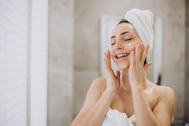 Mujer joven aplicando crema facial en la cara con una toalla en la cabeza