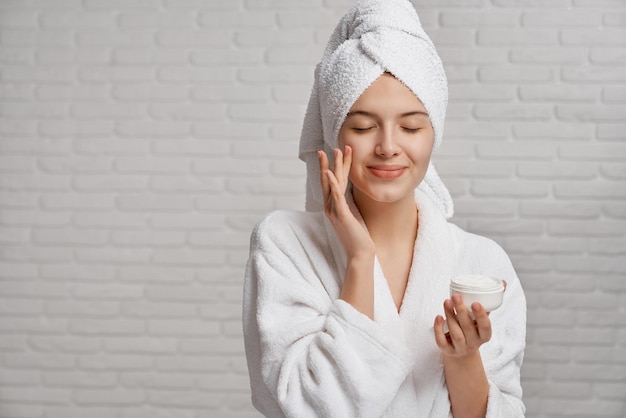 Mujer joven aplicando crema hidratante en la cara con los ojos cerrados