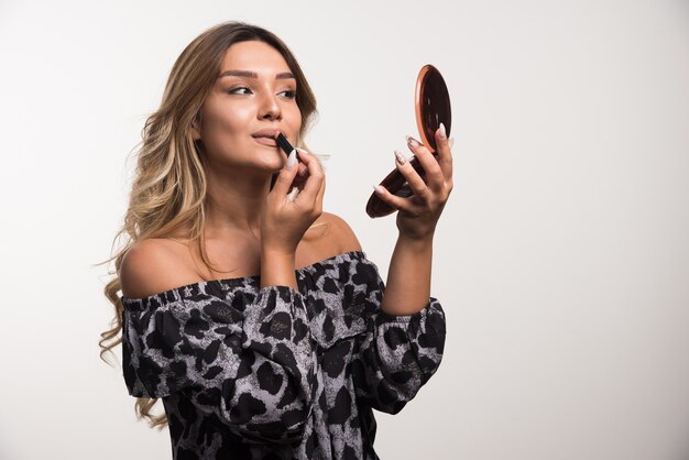 Mujer joven aplicando lápiz labial en la pared blanca.