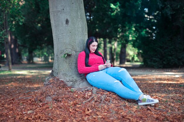 mujer joven apoyada contra un árbol usa un teléfono inteligente