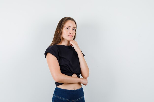 Mujer joven apoyando la barbilla en la mano en blusa negra, pantalones y aspecto sensible