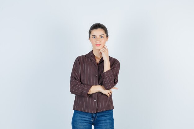 Mujer joven apoyando la barbilla en la mano con camisa a rayas, jeans y aspecto encantador. vista frontal.