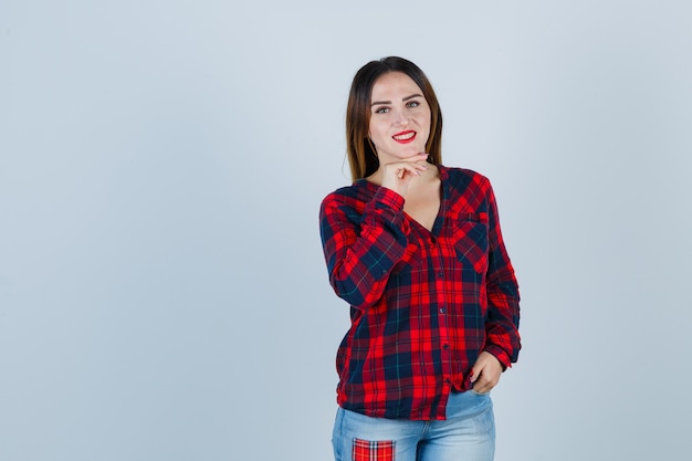 Mujer joven apoyando la barbilla en la mano, con la mano en el bolsillo en camisa a cuadros, jeans y mirando feliz. vista frontal.