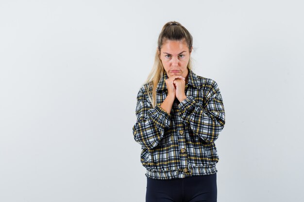 Mujer joven apoyando la barbilla en las manos entrelazadas en camisa, pantalones cortos y mirando pensativo. vista frontal.