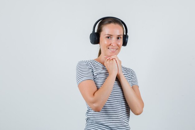 Mujer joven apoyando la barbilla en las manos entrelazadas en camiseta, auriculares y mirando feliz. vista frontal.