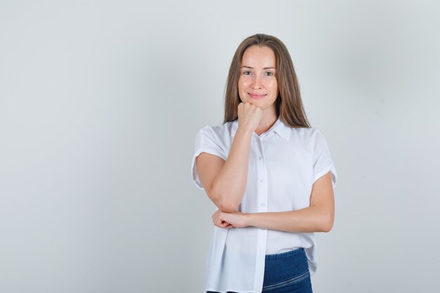 Mujer joven apoyando la barbilla en el puño en camiseta, jeans y mirando alegre