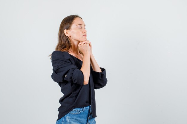 Mujer joven apoyando la barbilla en los puños con los ojos cerrados en camisa, pantalones cortos y mirando esperanzado. .