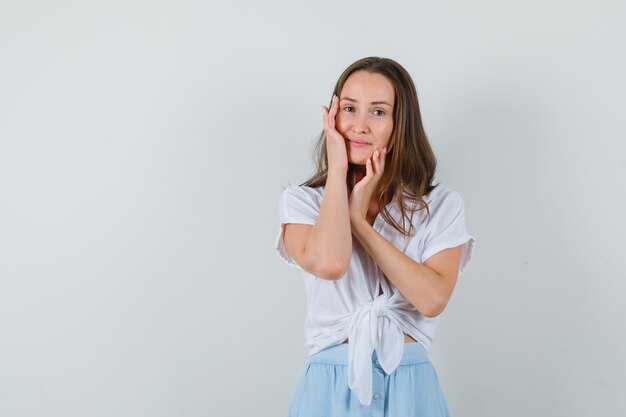 Mujer joven apoyando las mejillas en la palma de la mano en blusa blanca y falda azul claro y luciendo elegante