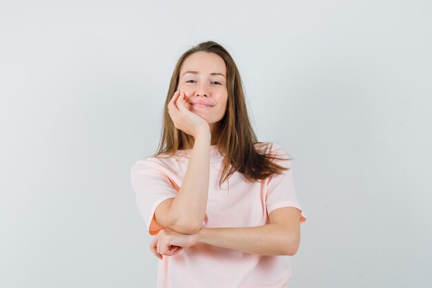 Mujer joven apoyando el mentón en la palma en camiseta rosa y mirando alegre.