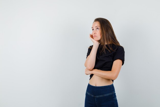 Mujer joven apoyando el mentón en la palma de la mano en blusa negra, pantalones y mirando pensativo.