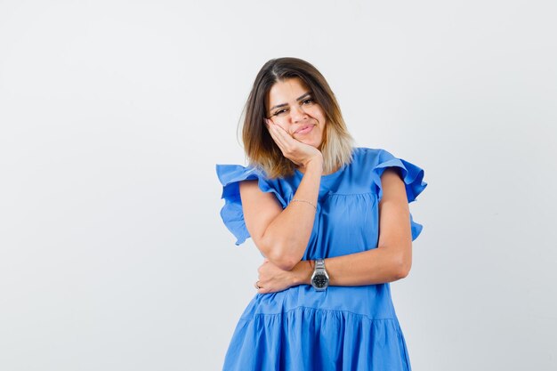 Mujer joven apoyándose en la mejilla en la palma levantada en vestido azul y mirando optimista