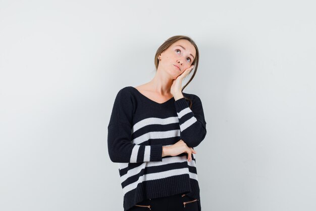 Mujer joven apoyándose la mejilla en la palma, de pie en pose de pensamiento en prendas de punto a rayas y pantalones negros y mirando pensativo