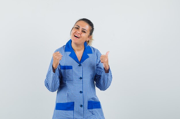 Mujer joven apretando el puño mientras muestra el pulgar hacia arriba en camisa de pijama de cuadros azules y mirando feliz. vista frontal.