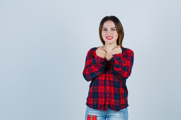 Mujer joven apretando los puños sobre el pecho en camisa a cuadros, jeans y mirando alegre, vista frontal.