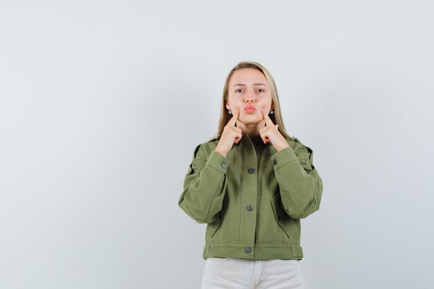 Mujer joven apretando sus mejillas en chaqueta verde, jeans y mirando enfocado. vista frontal.