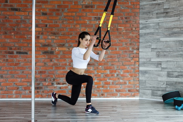 La mujer joven apta hace estiramientos, empuja hacia arriba y levanta los anillos de gimnasia en el gimnasio