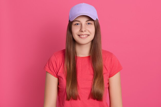 Mujer joven apta que lleva la camiseta casual que se coloca aislada sobre la pared rosada. Hermosa modelo en gorra de béisbol