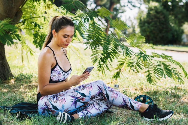 Mujer joven de la aptitud que se sienta en el jardín usando smartphone